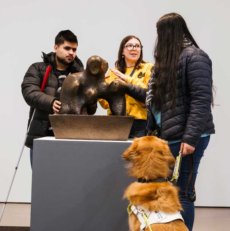 Two visitors and a Guide Dog with an Information Assistant interacting with Henry Moore's Mother and Child: Arch, 1959 in Beyond the Visual at Henry Moore Institute 28 November 2025 – 19 April 2026. Photo: Joanne Crawford.