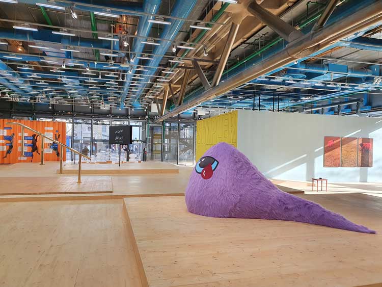 General exhibition view of Hassan Khan: Blind Ambition including: Purple Stuffed Creature with Bleeding Eye, 2019, foam, long hair fur fabric with digital print, 300 x 150 x 150 cm. Collection Centre Pompidou, Musee national d’art modern; Dense Object, 2013, metal object, wooden table, 10 x 5.20 cm. Collection of the artist; The Rams, 2006, three photographs printed on photographic paper and mounted on aluminium, 160 x 100 cm (each); Simpler Times, 2019, wall painting, 350 x 700 cm; Banque Bannister, 2010, brass, 209 x 206 x 22 cm; The Slapper and the Cap of Invisibility, 2015, black and white video, sound. 8 min 34 sec; Abstract Music, 2015, glass sculptures, wooden platform and plinths, variable dimensions. Museum für Moderne Kunst (MMK), Frankfurt. The acquisition was generously supported by the BHF Bank Stiftung; 2013 Curtain Remix, 2021, printed fabric, 350 x 840 cm. Photo courtesy of Hassan Khan.