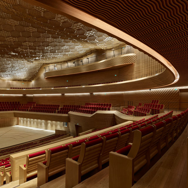 Shigeru Ban’s trademark cardboard tubes appear, upholstered and set into the wooden seat-frames and also in the ceiling: here, a honeycomb of 916 wood hexagons filled with 28,000 fireproofed paper tubes casting shadows across the space as the light changes. Photograph: Didier Boy de la Tour.