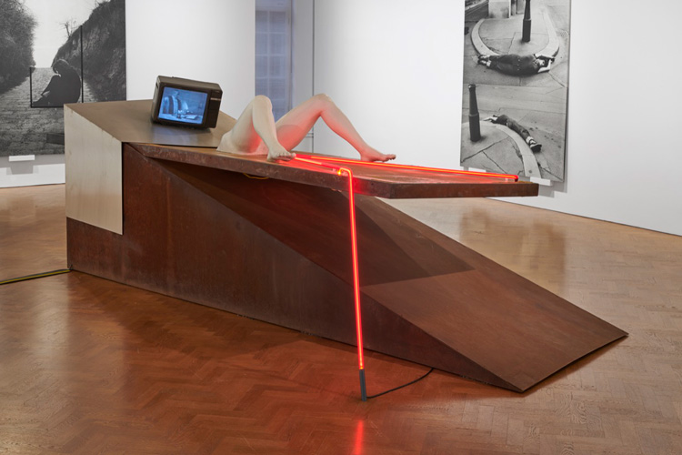 VALIE EXPORT. Geburtenbett, 1980. Wedge, rusted steel structure. Bed: steel frame with bedsprings covered with polyester; Women's legs: fibreglass reinforced with synthetic resin; 
Upholstery: stainless steel; Fluorescent tubes: red ruby glass. Video: sound, monitor, DVD, looped, 150 x 470 x 165 cm (59.06 x 185.04 x 64.96 in). © VALIE EXPORT/ Bildrecht 2019. Courtesy Galerie Thaddaeus Ropac, London | Paris | Salzburg. Photo: Ben Westoby.